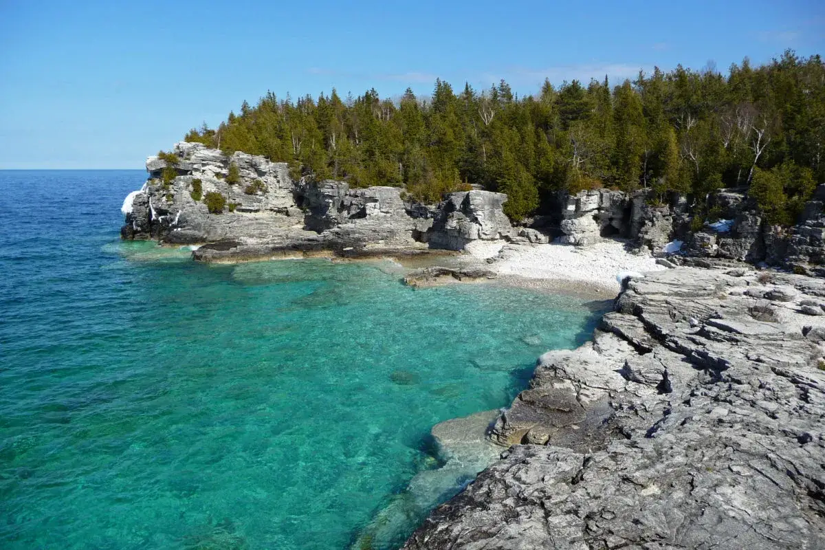 teal blue water by a rocky shoreline with forest | ontario beaches